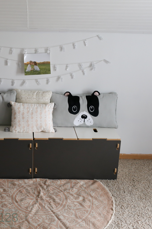 Toy storage boxes used as a reading bench with added pillows and string lights for hanging pictures.