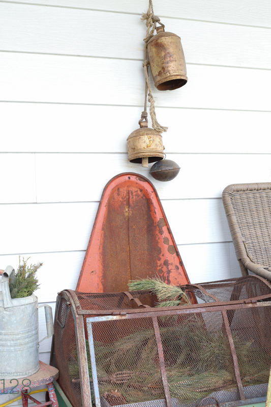 Farmhouse Christmas front porch decorating. Old red sled and brass bells with the pine branch filled greenhouse and watering can.