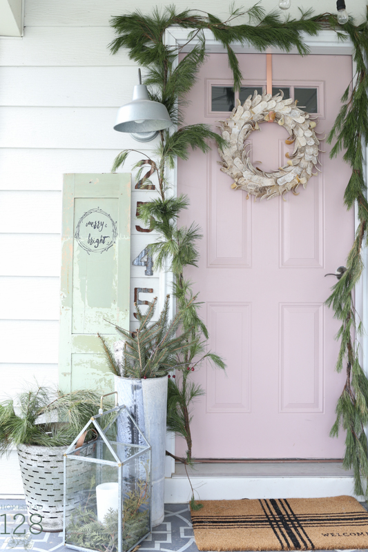 Farmhouse Christmas front porch decorating. A pink door is decorated with loads of garland and pine branches, a vintage door, and galvanized tin.