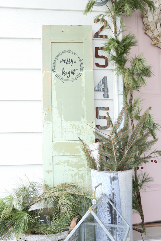 Farmhouse Christmas front porch decorating. A pink door is decorated with loads of garland and pine branches, a vintage door, and galvanized tin.