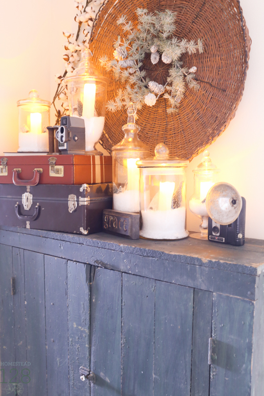 Farmhouse Christmas decorating. Old cupboard is decorated with small suitcases, and glass jars filled with candles, faux snow, and snow globe decor.
