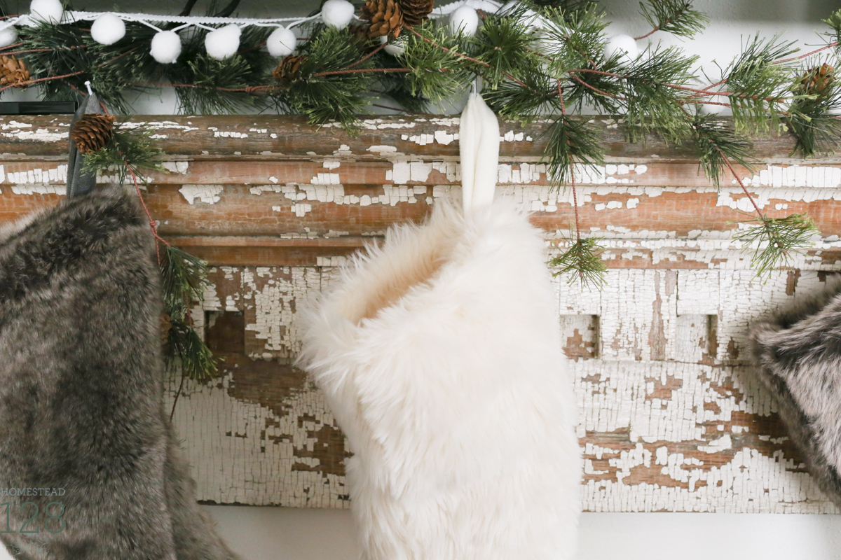 Farmhouse Christmas decorating. Furry and neutral stockings hung on a chippy white mantle with pine tree garland.